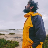 Mand med skaeg i gul og morkeblaa kystnaer komfort jakke kigger op mod himlen ved kysten