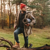 Mand med roed hue, jaegerens skjold jakke og sovepose staar i skov